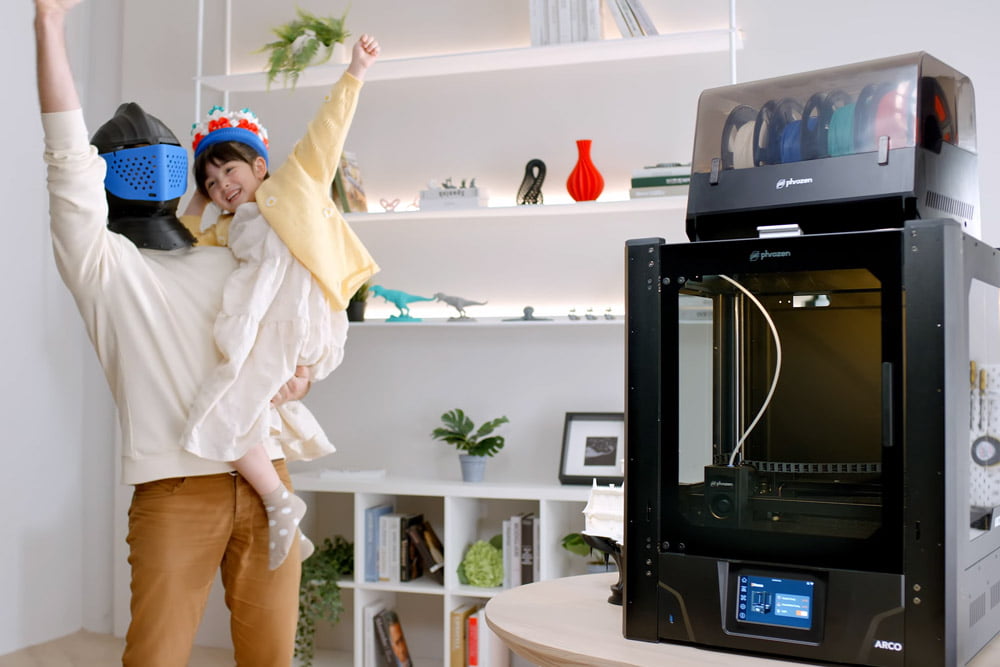 Adult lifting a child wearing a 3D printed crown, with Phrozen Arco printer in background.
