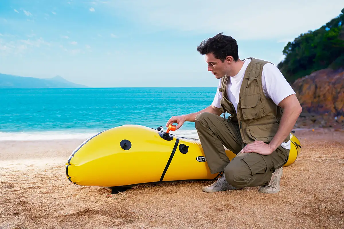 Person using ATOMIC PUMP to inflate a yellow inflatable kayak on a sandy beach, with a scenic ocean backdrop.